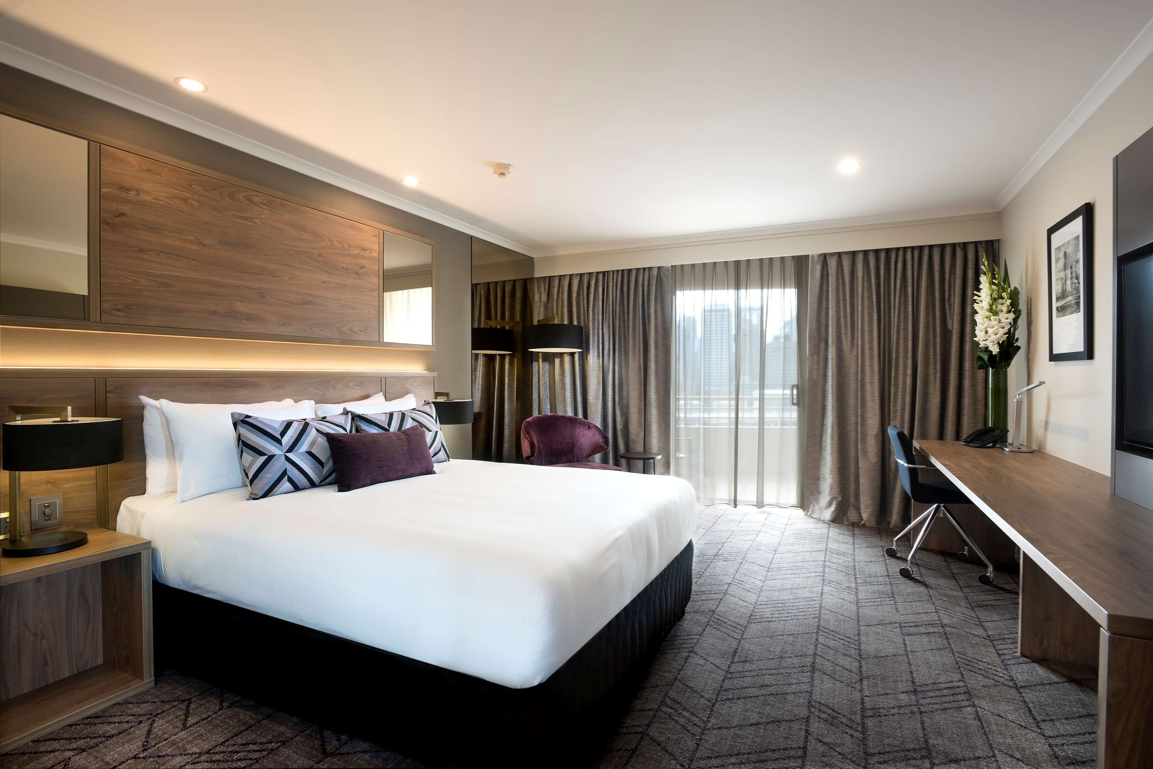 Een hotelkamer met een groot bed, een bureau, een stoel, een lamp en een groot raam met gordijnen.