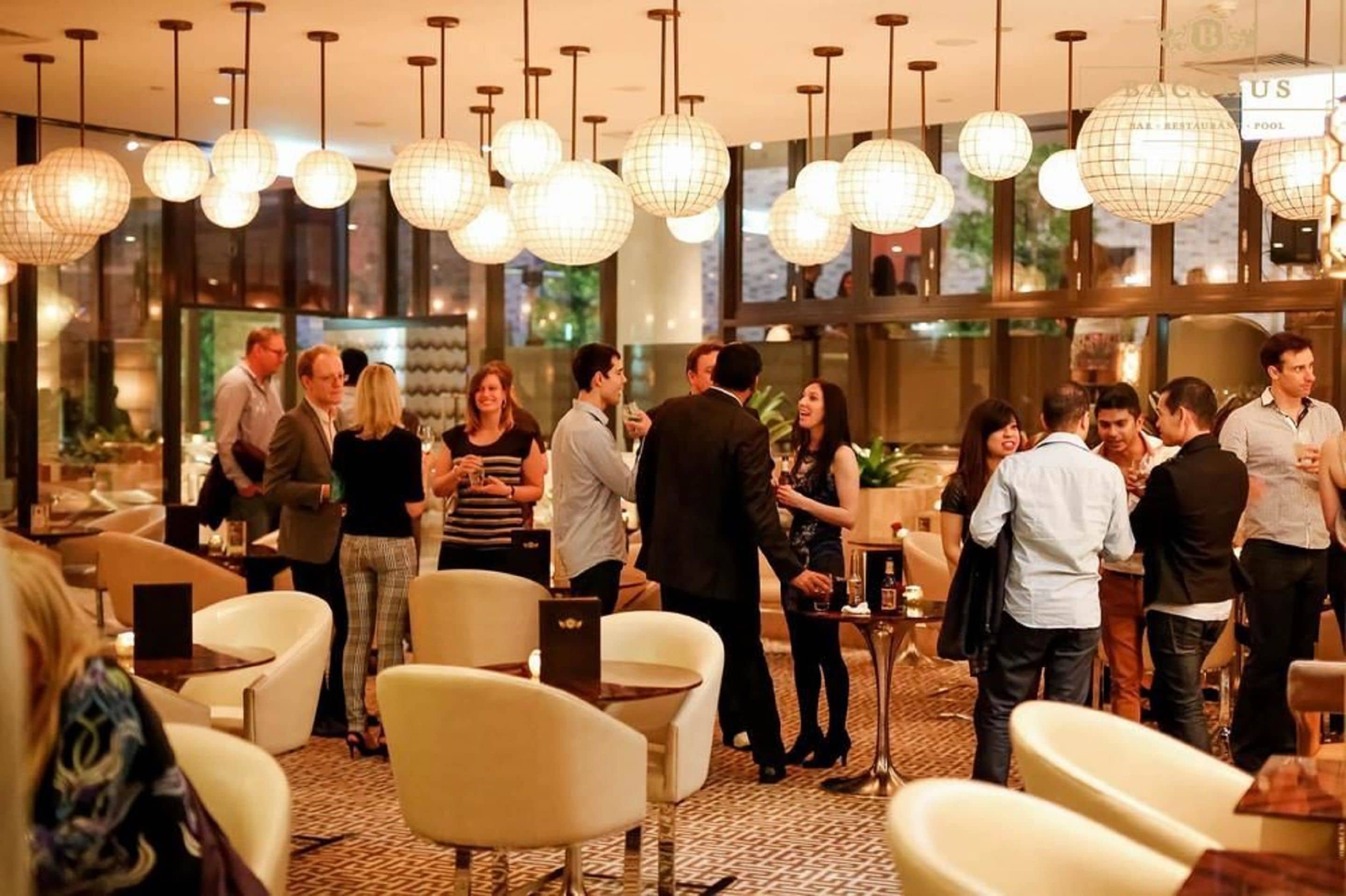 Un encuentro social en un moderno vestíbulo de hotel, con lámparas esféricas colgantes, asientos cómodos y un cartel que indica zona de restaurante y piscina.