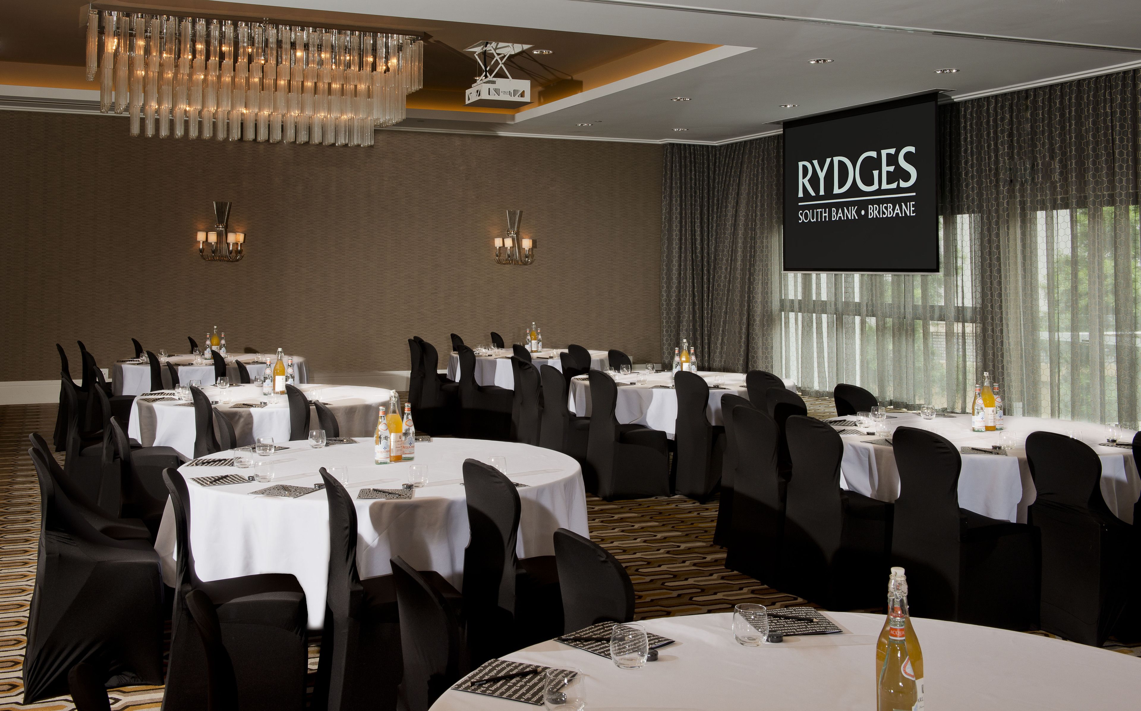 A conference room at Rydges South Bank, Brisbane with multiple round tables, black chairs, and a large screen displaying the hotel name.