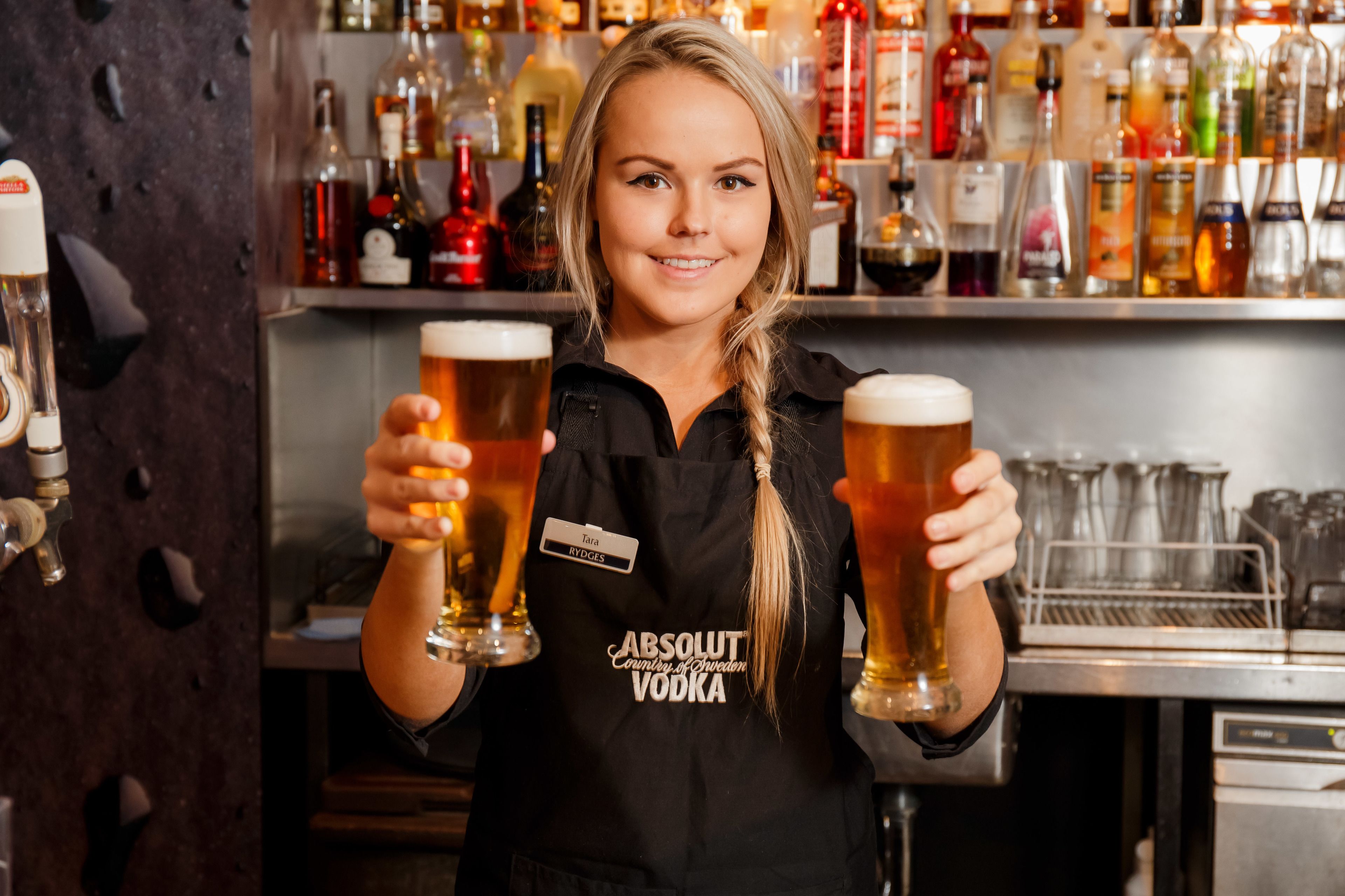 Un cantinero sostiene dos vasos de cerveza en un bar con una variedad de botellas al fondo.