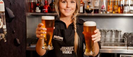 A bartender holding two glasses of beer in a bar with a variety of bottles in the background.