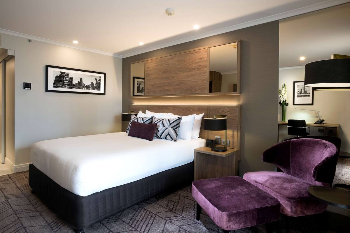 A modern hotel room with a large bed, a vanity with a mirror, a purple armchair, and a small table.
