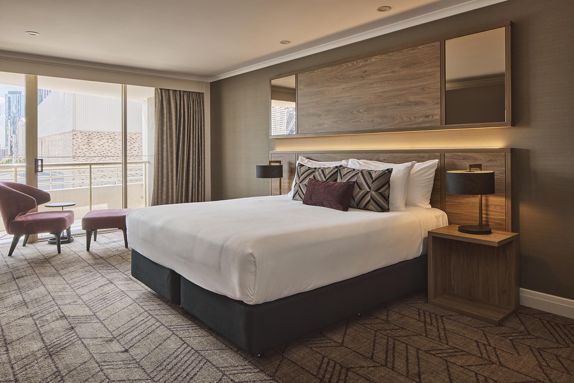 Habitación de hotel con una cama grande, un escritorio, una silla y vistas de la ciudad a través de amplios ventanales.
