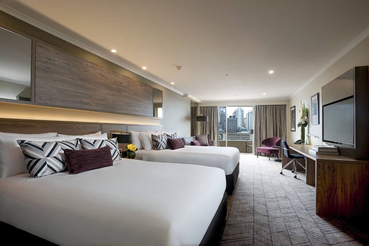 A hotel room with two beds, a desk, a chair, a TV, and a view of the city.