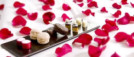 A dessert platter with macarons, chocolate, and small cakes, surrounded by rose petals on a white surface.