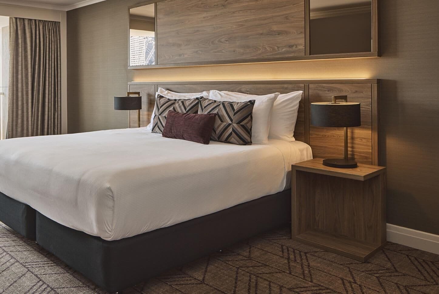 A hotel room with a large bed, a nightstand with a lamp, and a view of the city through a window.