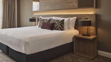 A hotel room with a large bed, a nightstand with a lamp, and a view of the city through a window.