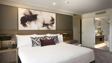 Una habitación de hotel moderna con una cama grande, una mesita de noche con teléfono, una obra de arte colgada en la pared y un baño visible a través de una puerta abierta.