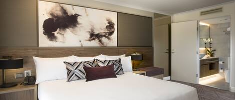 A modern hotel room with a large bed, a nightstand with a phone, a wall-mounted artwork, and a bathroom visible through an open door.