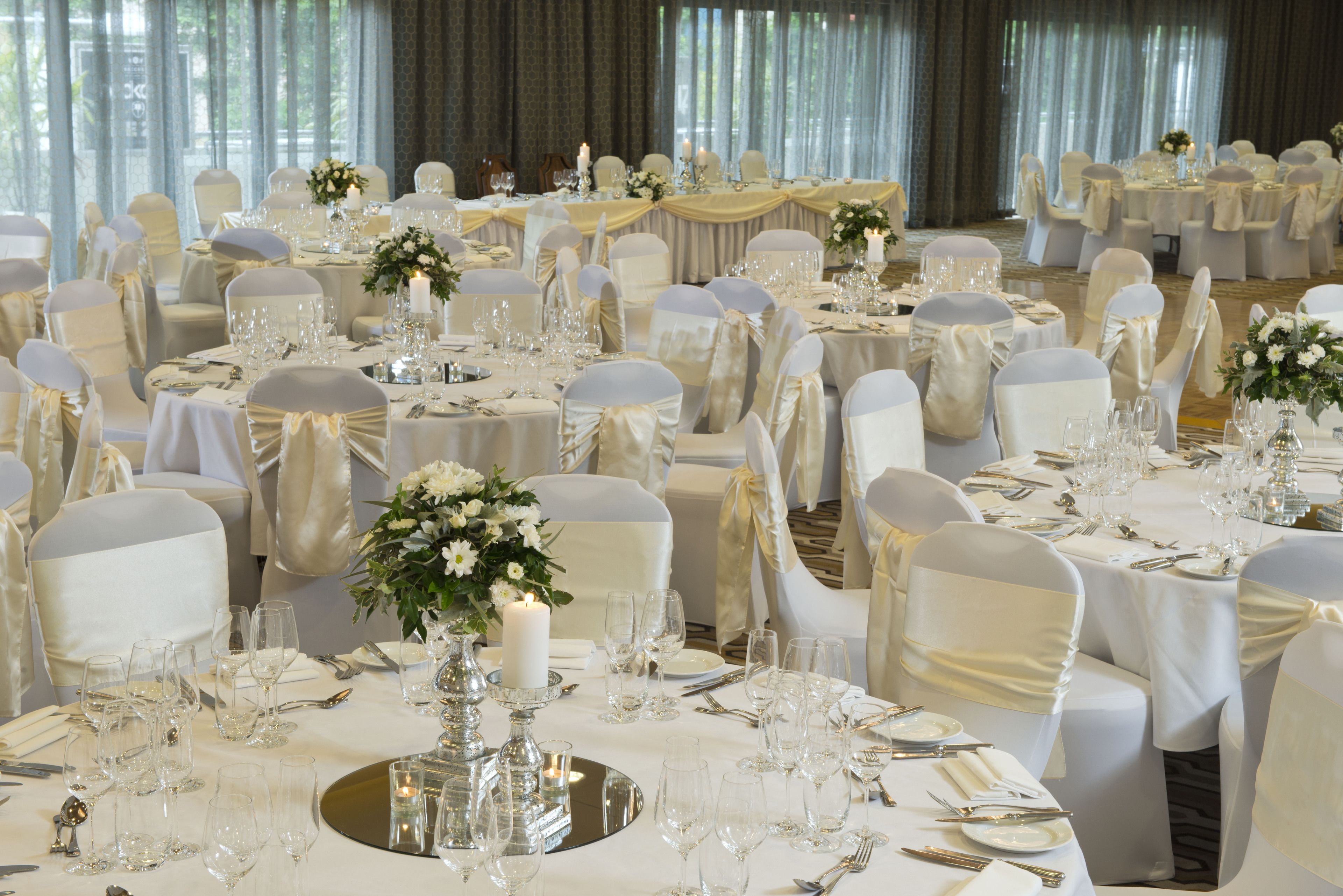 Un salón de banquetes con mesas redondas dispuestas para un evento formal, cada una con manteles blancos, cristalería y centros de mesa florales.