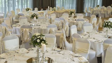 Un salón de banquetes con mesas redondas dispuestas para un evento formal, cada una con manteles blancos, cristalería y centros de mesa florales.