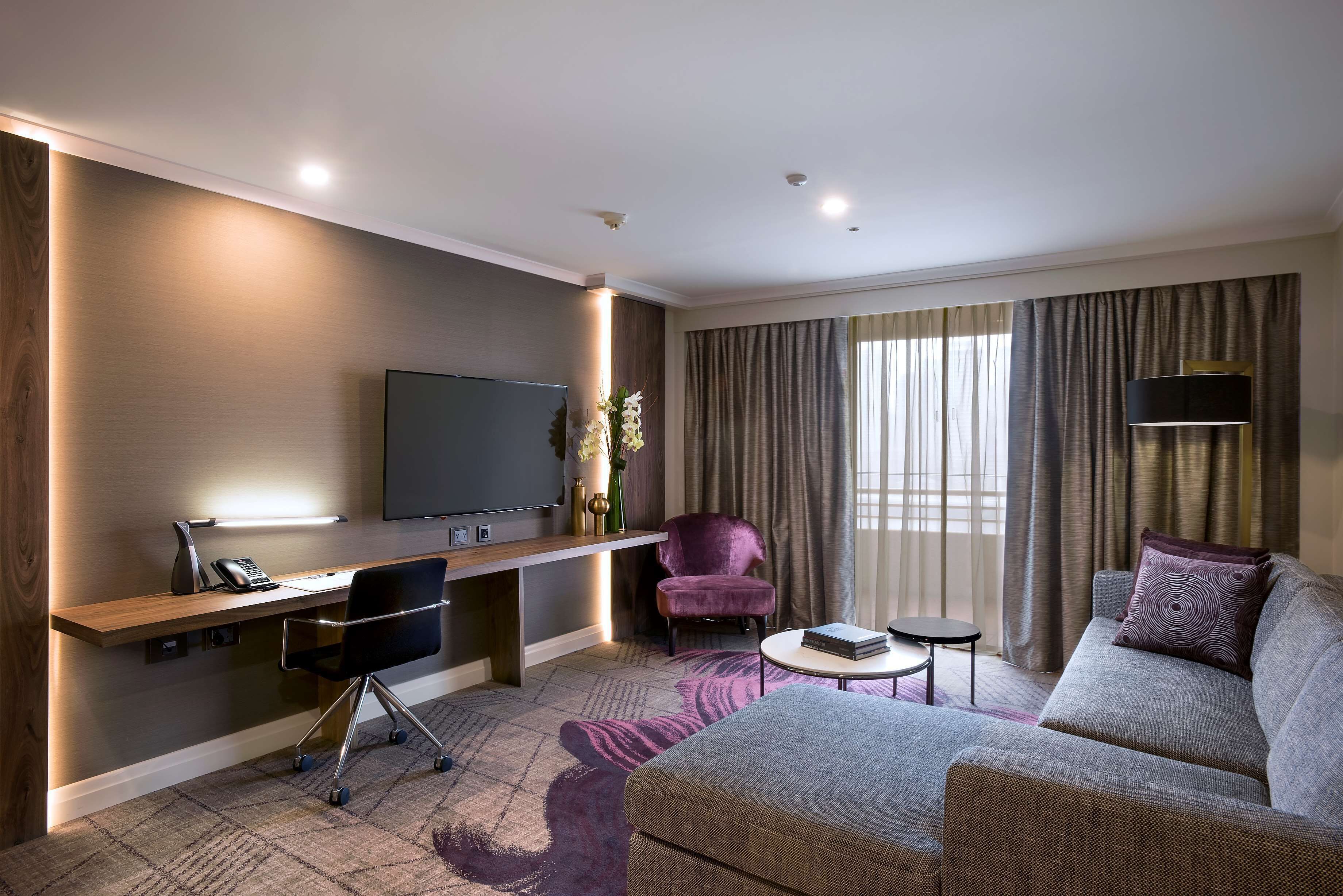 Una habitación de hotel moderna con televisión de pantalla plana, un escritorio con silla, un sofá y una mesita redonda.