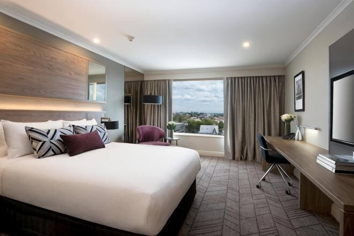 A hotel room with a large bed, a desk, a chair, a TV, and a view of the city.