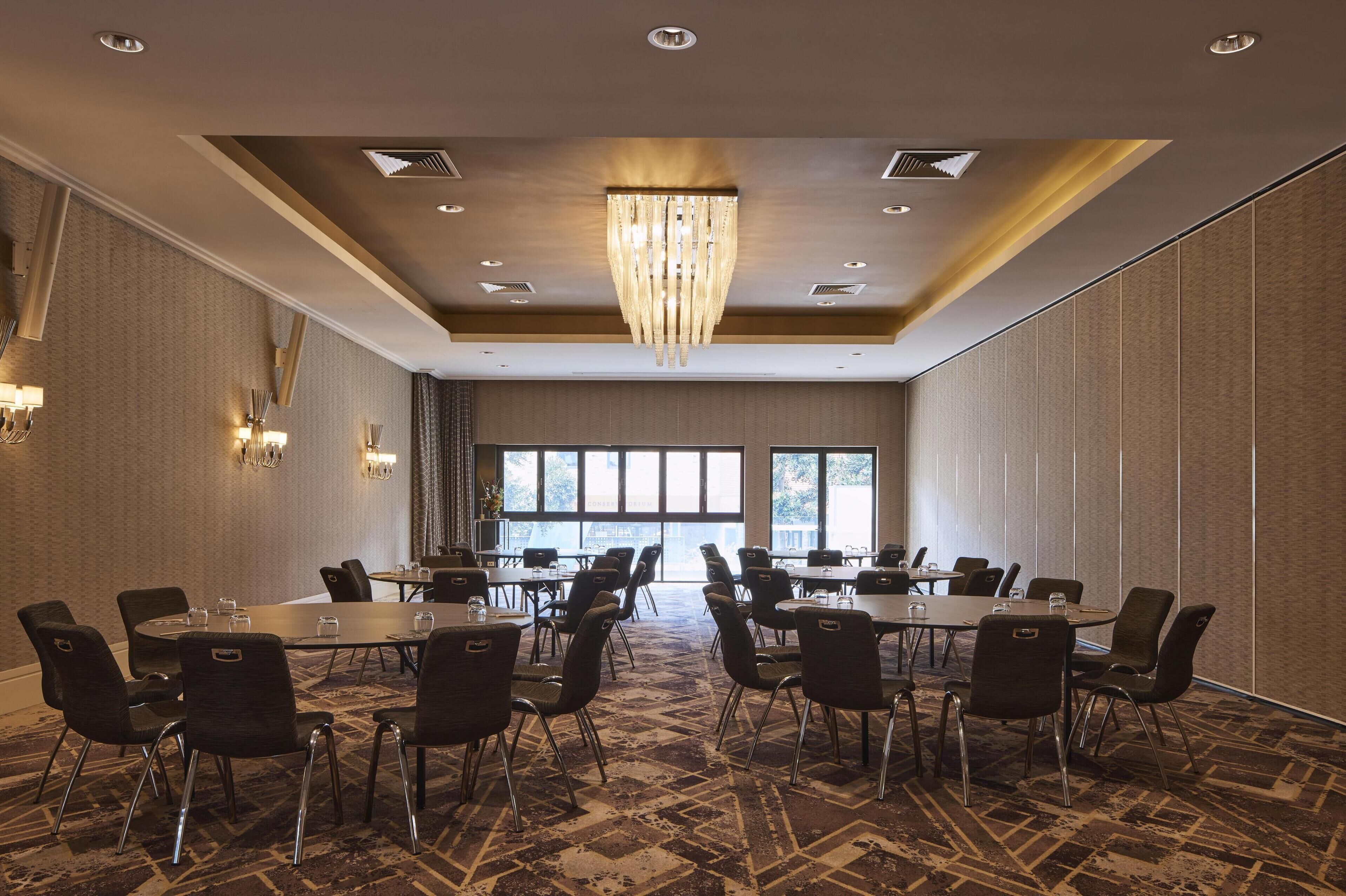 Un salón de conferencias con varias mesas y sillas, ventanas amplias y una araña de cristal.