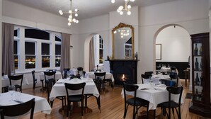 Un comedor con varias mesas y sillas, chimenea y ventanas grandes con cortinas.