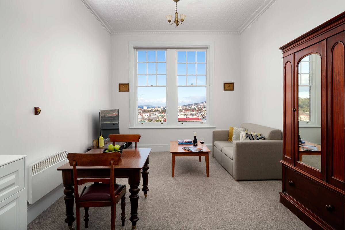 A living room with a dining table, chairs, a sofa, a cabinet, and a large window offering a city view.