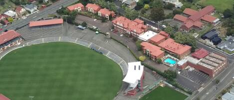 Vista aérea de un estadio con un amplio campo verde, rodeado de edificios residenciales y un área de estacionamiento.