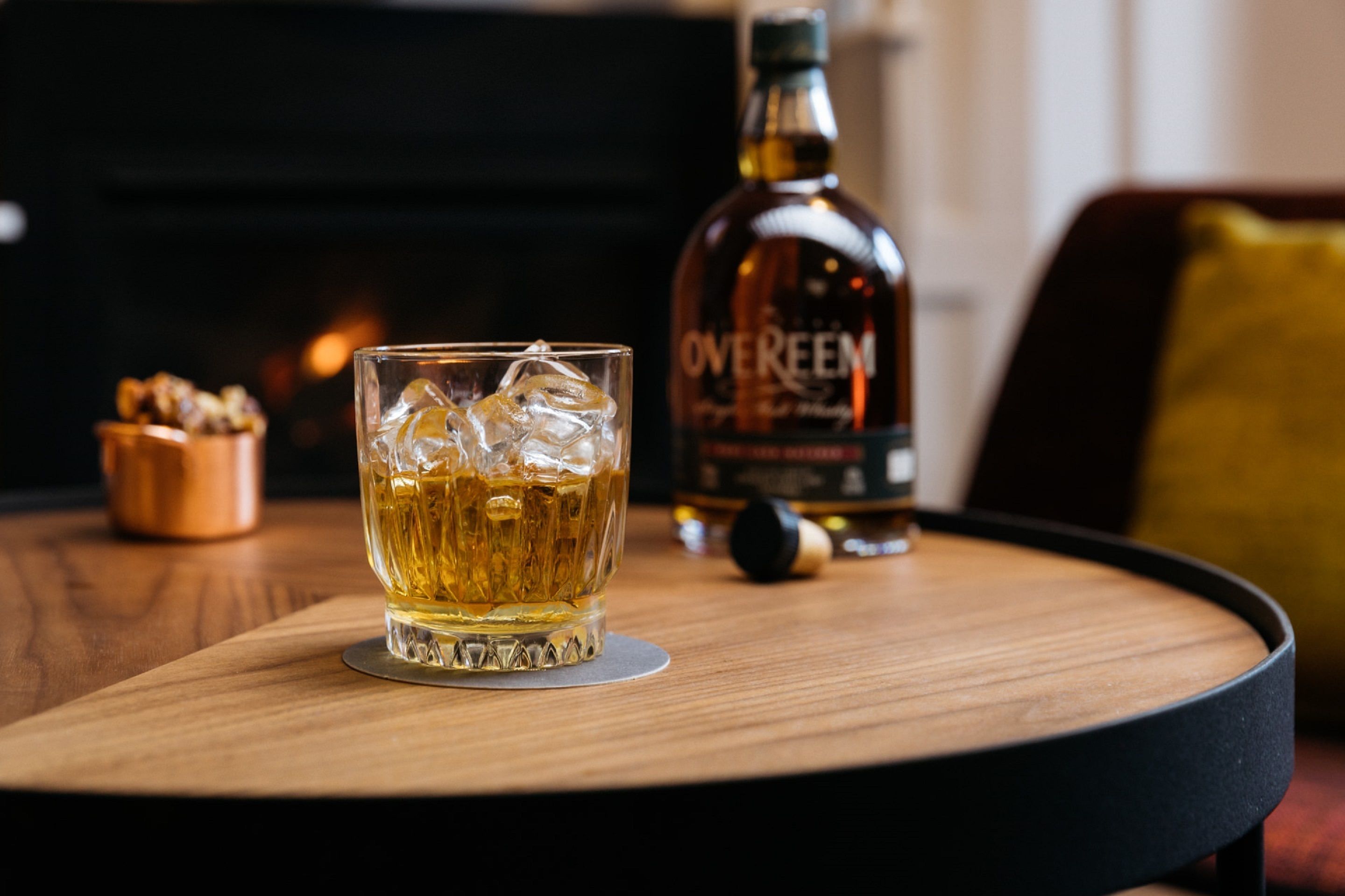 Ett glas whisky med isbitar på en underlägg av trä, en flaska Overeon-whisky och en liten skål med nötter på ett runt bord.