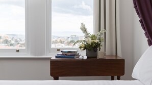 Un dormitorio con vistas a un paisaje urbano a través de amplios ventanales, una mesita de noche con libros y un florero, y una cama con ropa blanca.