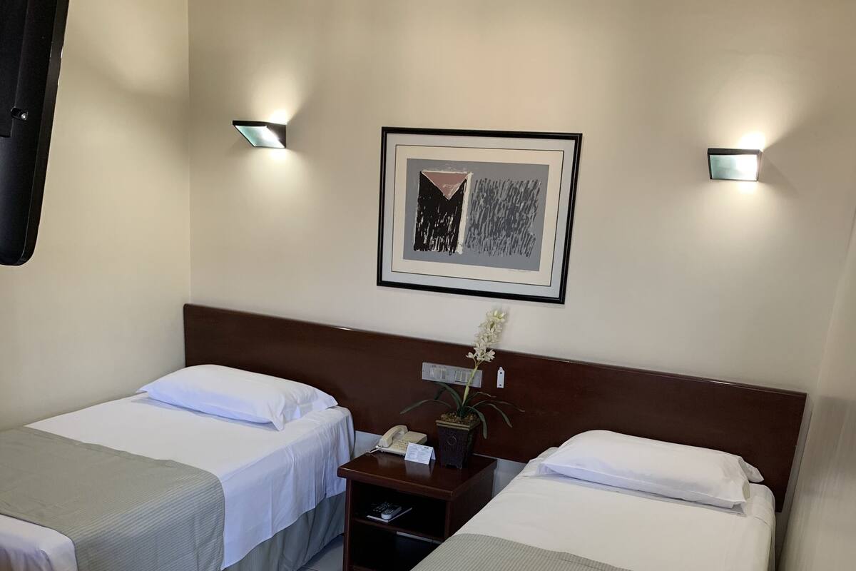 Une chambre d’hôtel avec deux lits simples, une table de chevet avec un téléphone et une petite plante, un tableau encadré au mur et un sol carrelé.