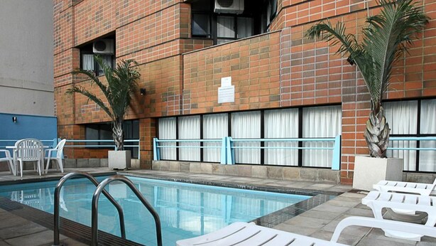 A swimming pool area with lounge chairs and a brick building in the background. - Cheverny Apart Hotel (Belo Horizonte)