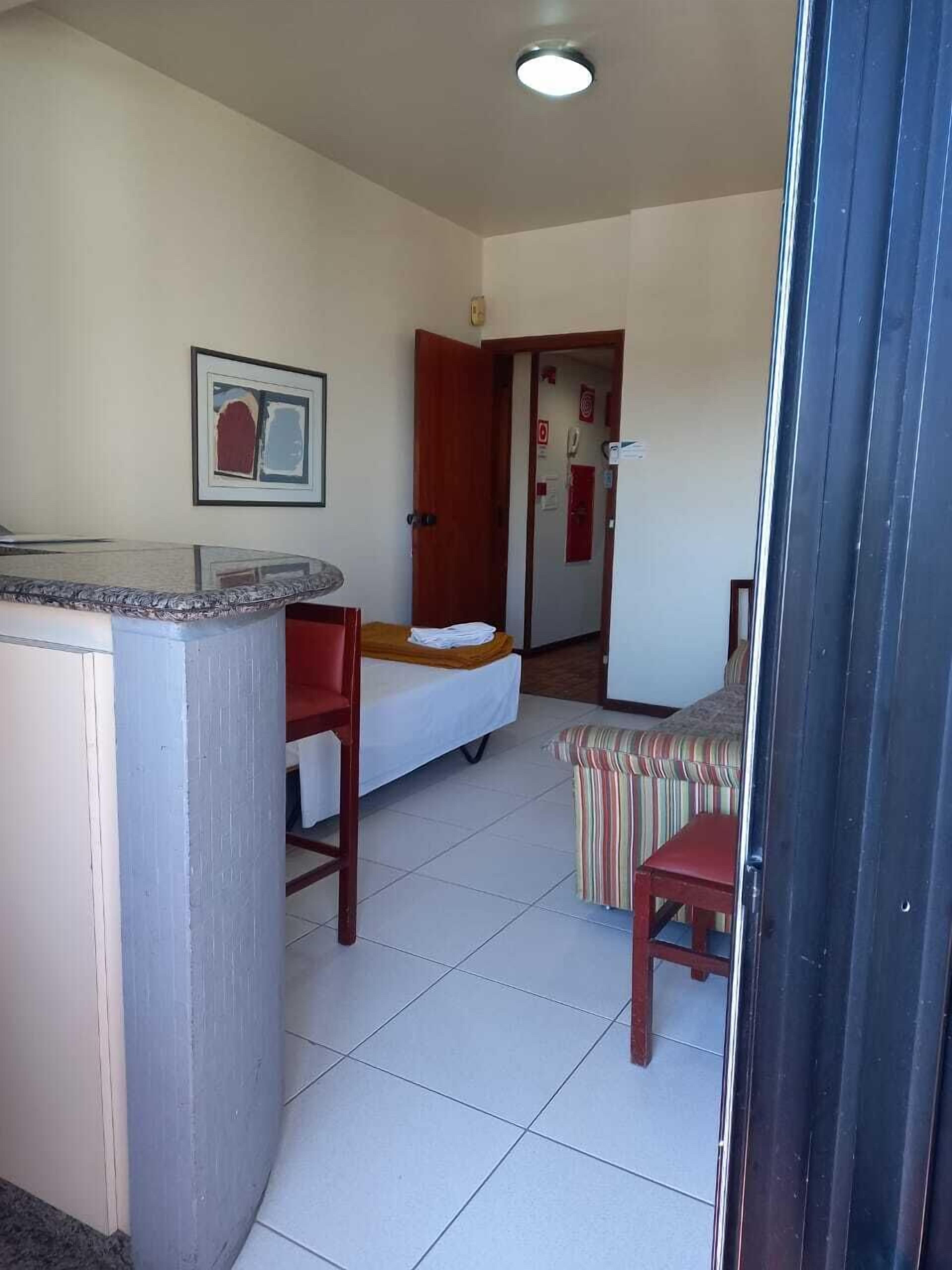 A compact hotel room with a kitchenette, a bed, a small table, and a striped chair.