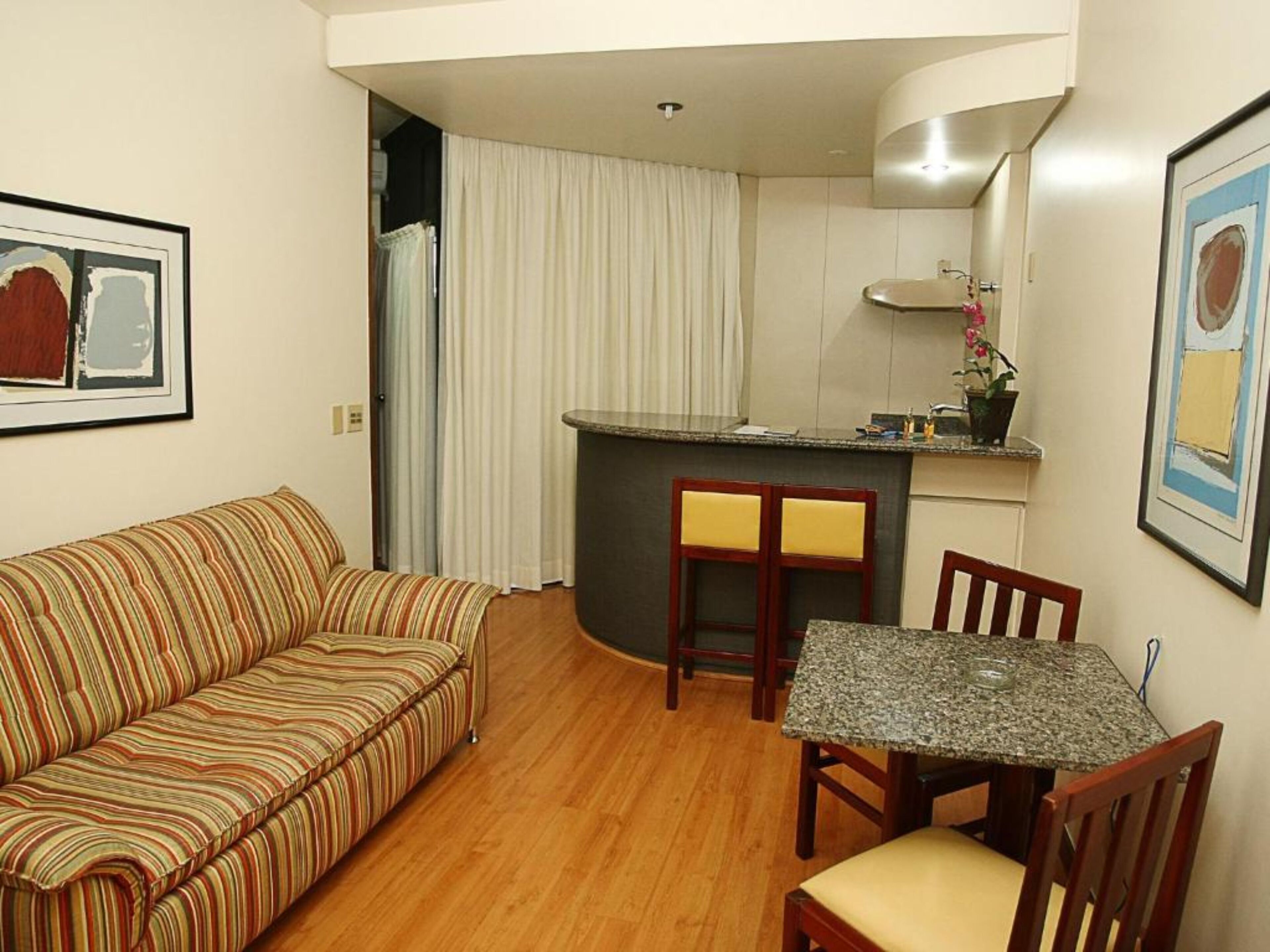 A compact living space with a striped sofa, a dining table with chairs, a kitchen area with a sink and countertop, and a window with curtains.