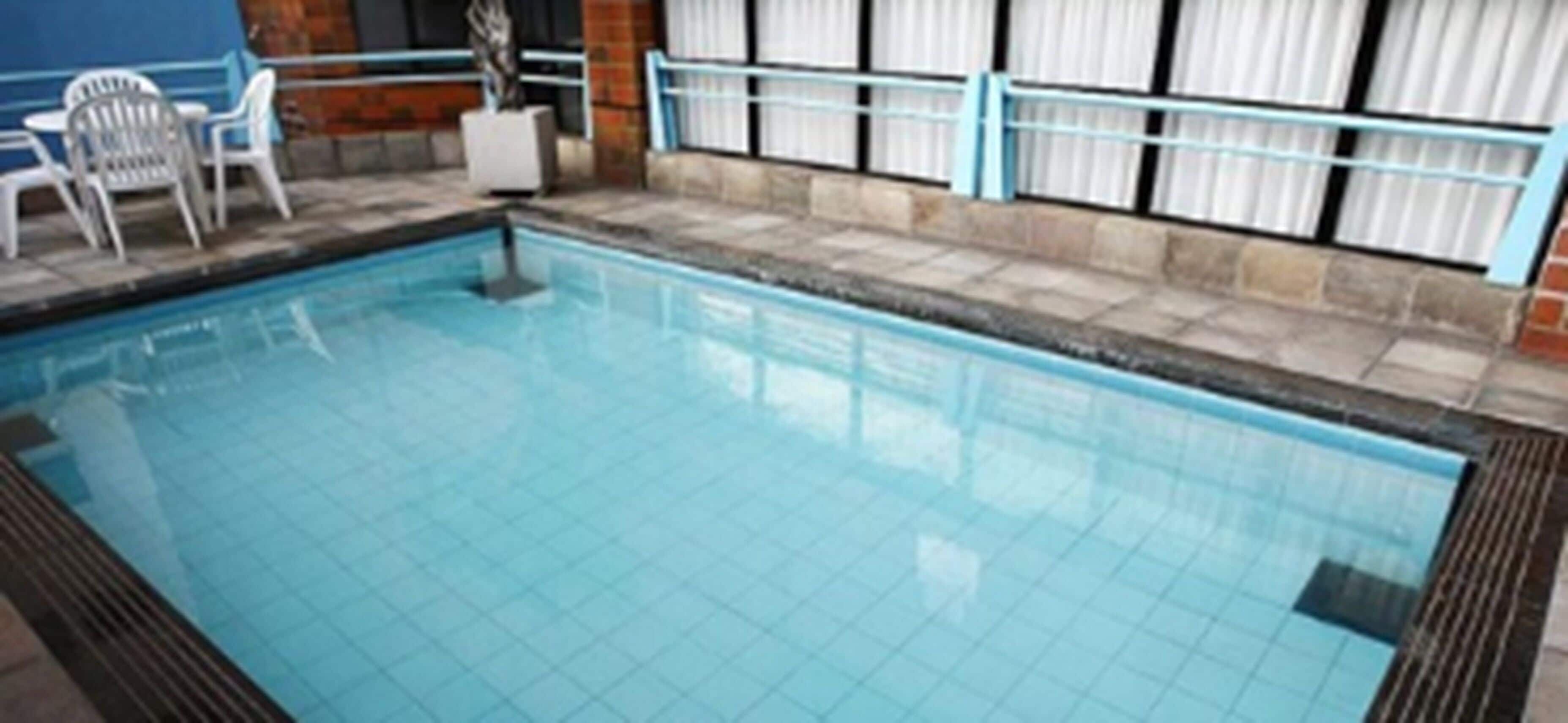 Una piscina con suelo de azulejos, un borde azul y una barandilla blanca. Hay sillas y una mesa blancas cerca de la piscina.