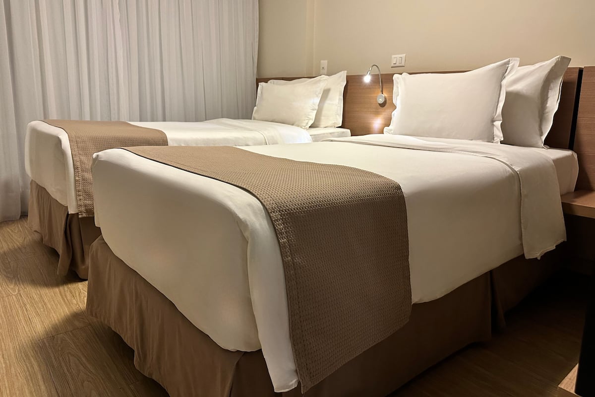 Un lit double avec une tête de lit en bois, une table de chevet avec une lampe et une fenêtre avec des rideaux blancs.