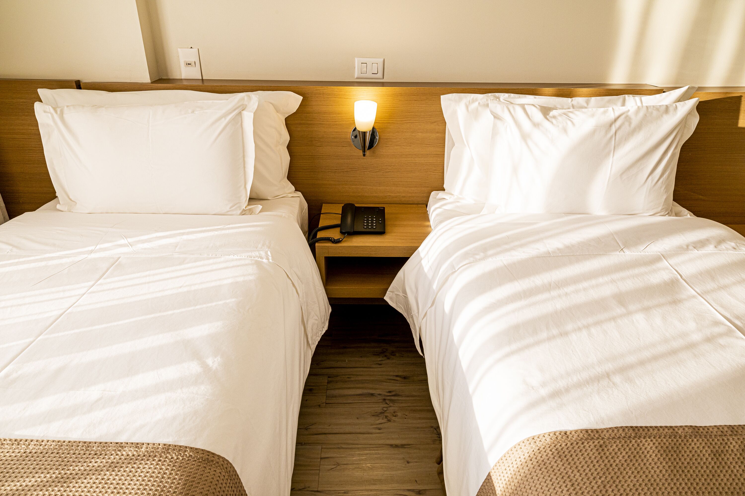 Quarto de hotel com duas camas de solteiro, cabeceira de madeira, mesa de cabeceira com abajur e telefone.
