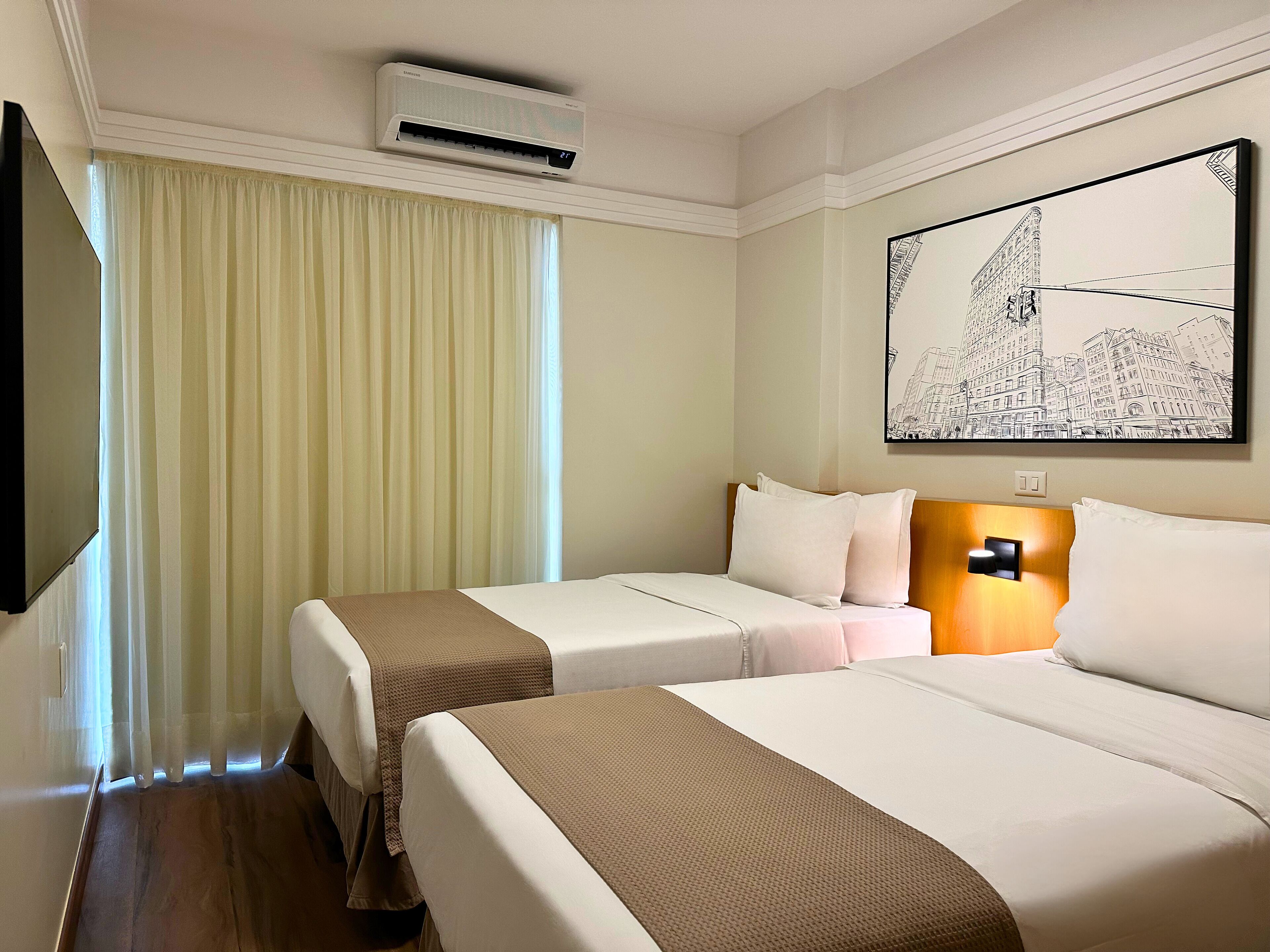 Quarto de hotel com duas camas, ar condicionado fixado na parede, uma janela grande com cortinas e uma obra de arte pendurada na parede.