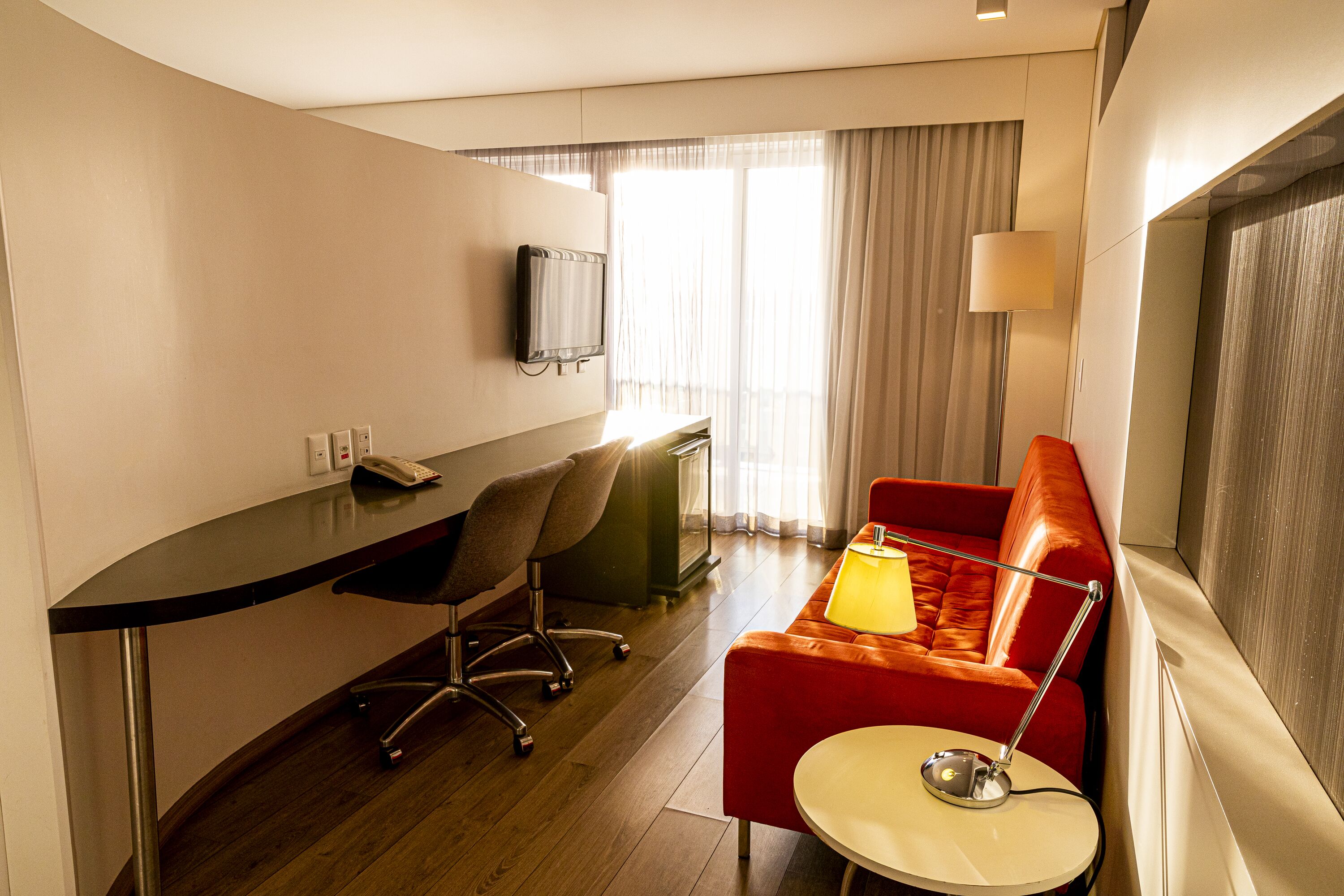 Une chambre d’hôtel avec un bureau, un canapé rouge, une petite table ronde, un téléviseur fixé au mur et une fenêtre avec des rideaux.