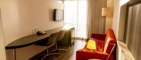 A hotel room with a desk, a red sofa, a small round table, a TV mounted on the wall, and a window with curtains.
