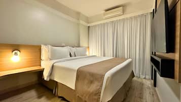 A hotel room with a bed, a bedside table with a lamp, a flat-screen TV, and an air conditioning unit.