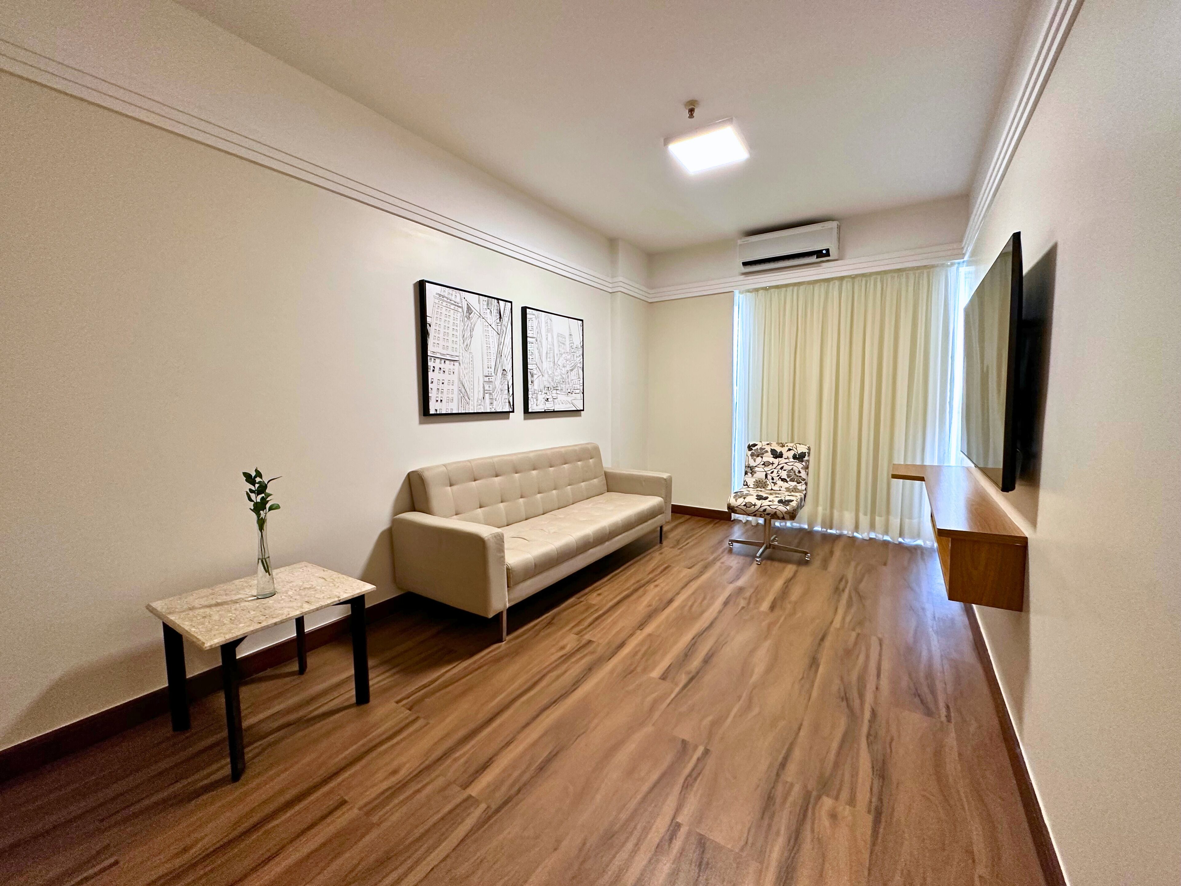 Una sala moderna con un sofá color beige, una mesita con un jarrón, y una silla estampada con flores. Se observan dos cuadros en la pared, una lámpara de techo y un equipo de aire acondicionado.