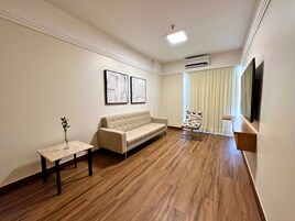Una sala moderna con un sofá color beige, una mesita con un jarrón, y una silla estampada con flores. Se observan dos cuadros en la pared, una lámpara de techo y un equipo de aire acondicionado.