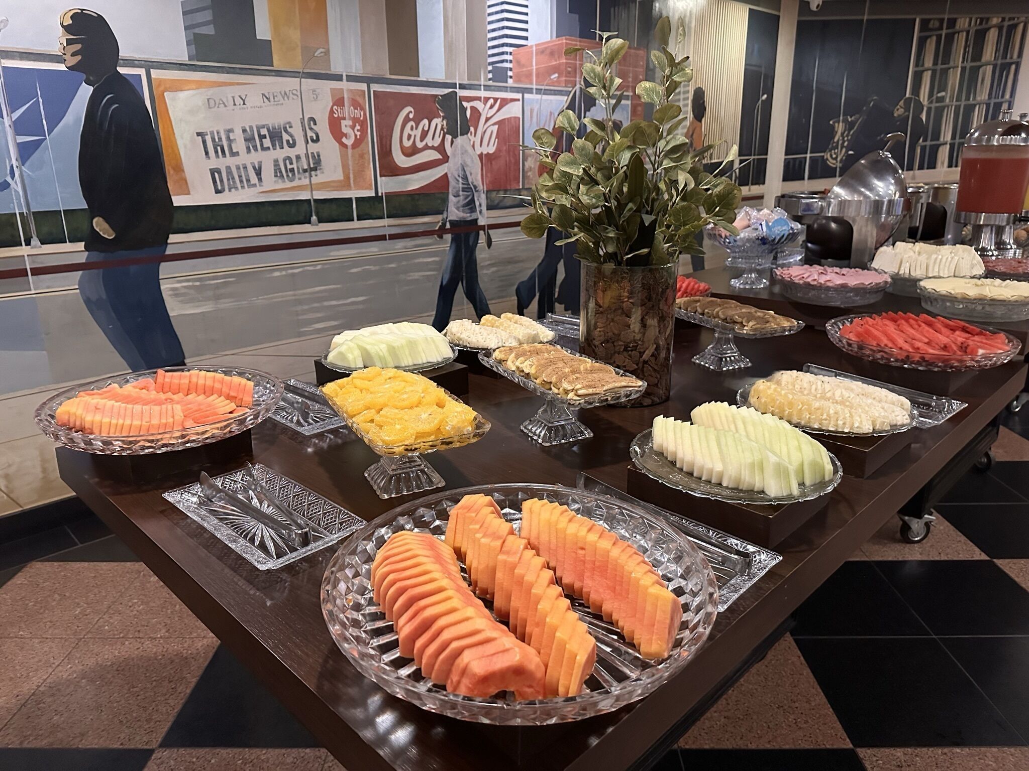 A buffet table with various food items, including sliced fruits and vegetables, in a public area with a Coca-Cola advertisement in the background.