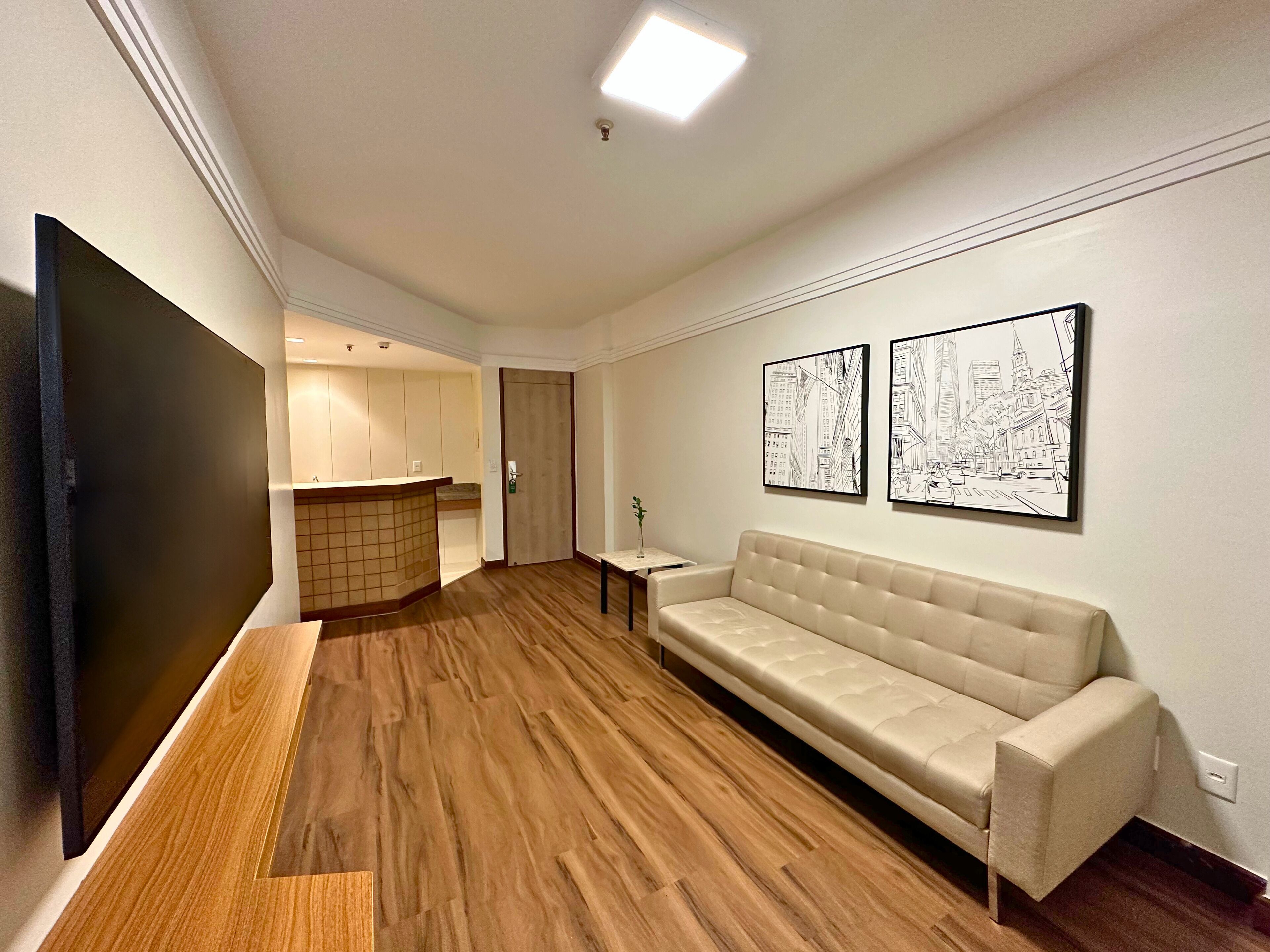 Uma sala de estar moderna com uma televisão de tela plana grande, um sofá bege, piso de madeira e duas obras de arte com tema urbano.