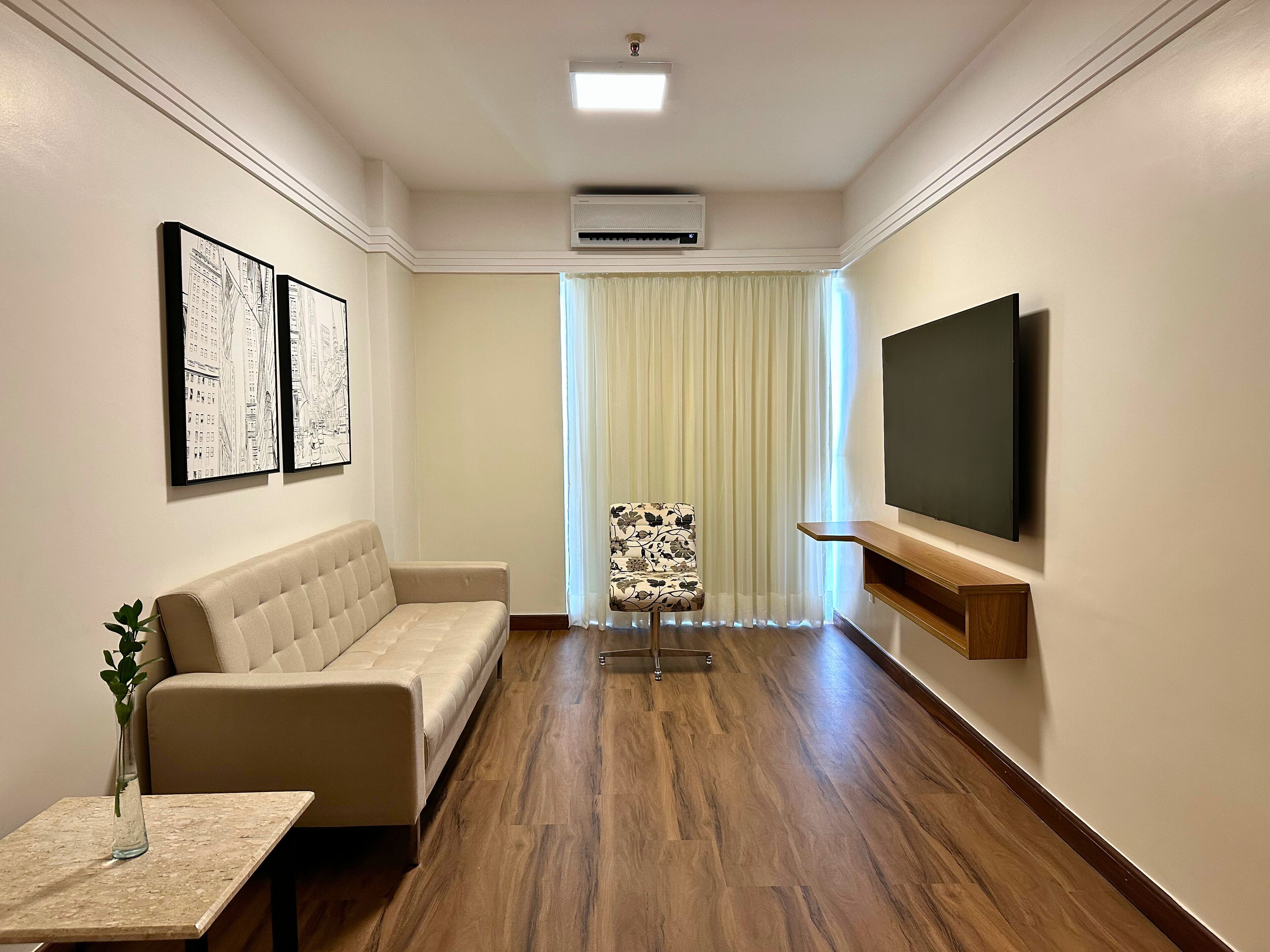 Quarto de hotel moderno com sofá, mesinha com um vaso, cadeira, TV fixada na parede e ar condicionado.