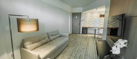 Uma sala de estar moderna com um sofá, uma luminária de chão e uma pequena mesa. Há uma TV fixada na parede e uma planta decorativa ao lado.