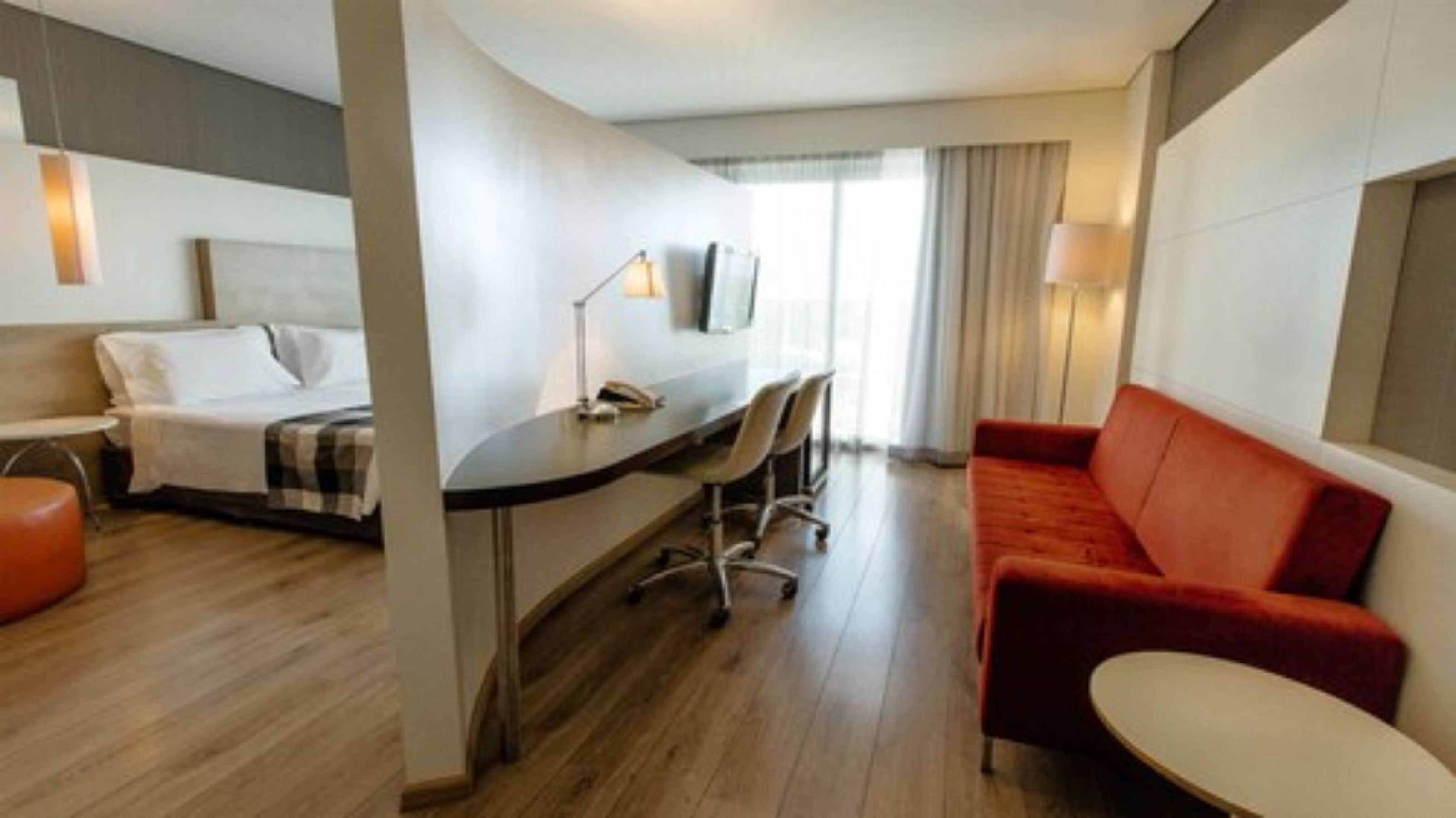 Una habitación de hotel con cama, escritorio y silla, sofá rojo y una mesita.