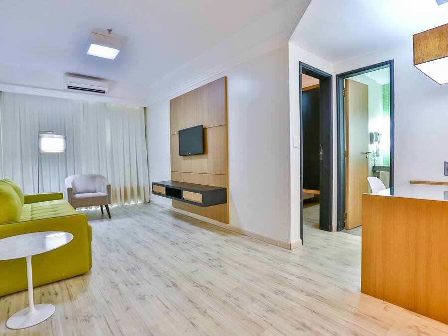 Una habitación de hotel moderna con un sofá verde, una pequeña mesa redonda, un televisor de pantalla plana montado en un panel de madera y un mostrador de recepción.