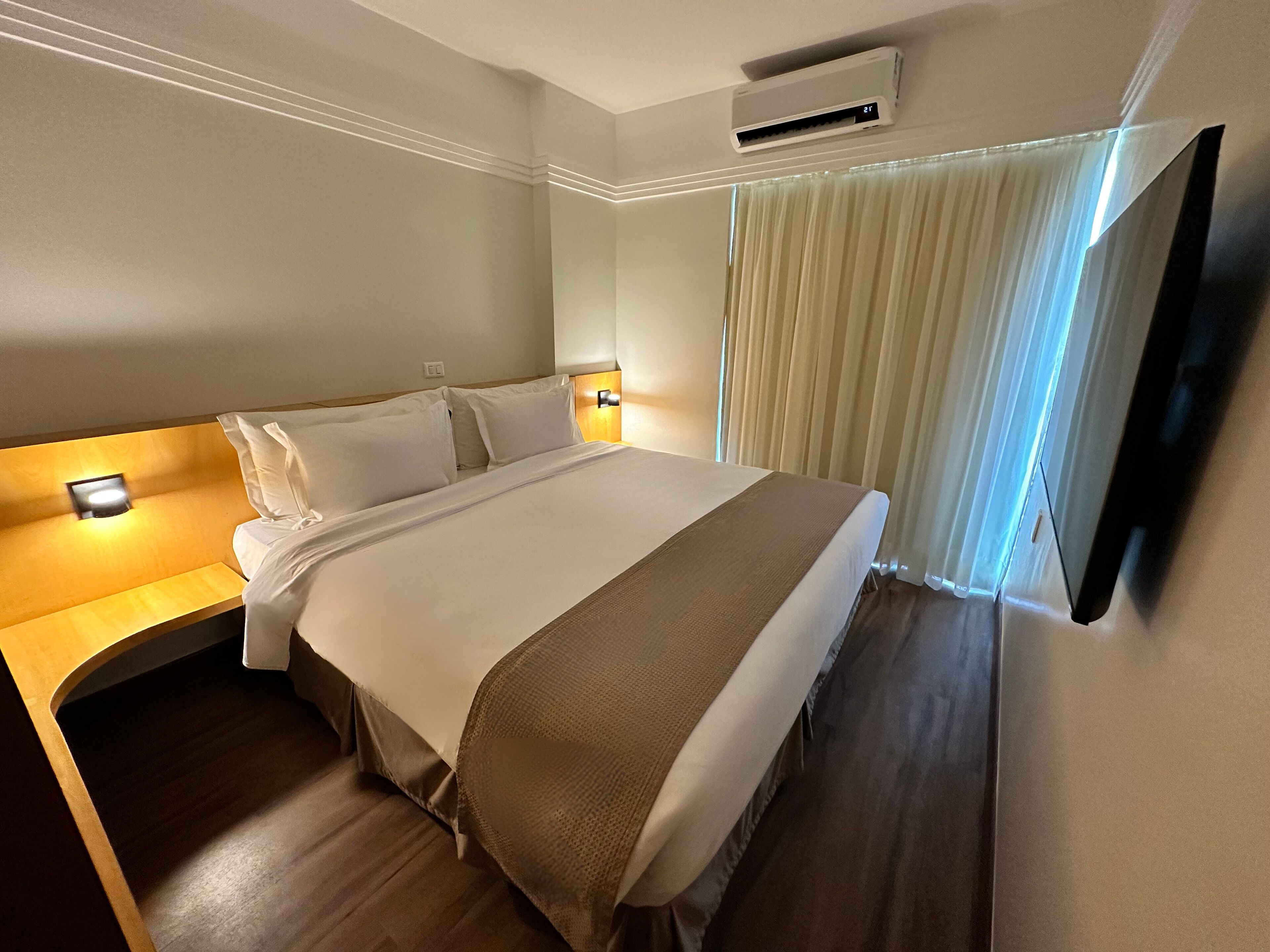 Habitación de hotel con cama, lámparas de noche, aire acondicionado, televisor y ventana con cortinas.