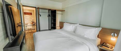 Quarto de hotel com uma cama grande, mesa de cabeceira com abajur, guarda-roupa e espelho.