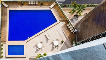 Una piscina en la azotea con área de azulejos azules, rodeada de sillas de descanso y mesas, y un pequeño jardín con plantas.
