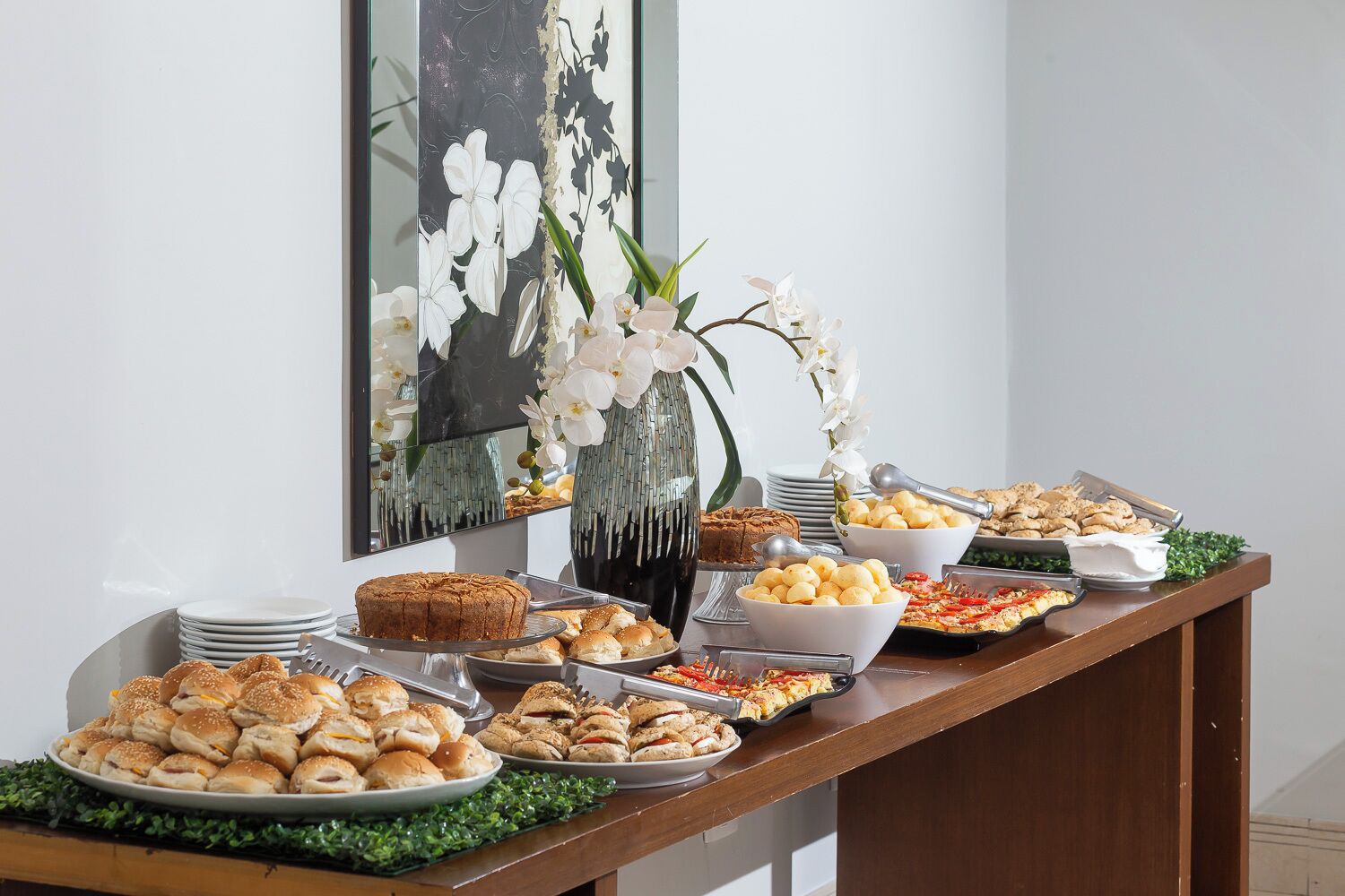 Une table de buffet proposant divers plats, notamment des pâtisseries, des sandwichs et un gâteau, placée devant un mur blanc orné d’une œuvre d’art encadrée et d’un vase de fleurs blanches.