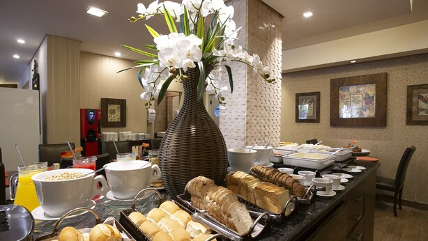 A buffet with various food items, a large vase with white flowers, and a coffee station. - Roochelle Hotel by Nobile (Curitiba)