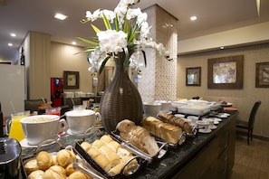 A buffet with various food items, a large vase with white flowers, and a coffee station. - Roochelle Hotel by Nobile (Curitiba)