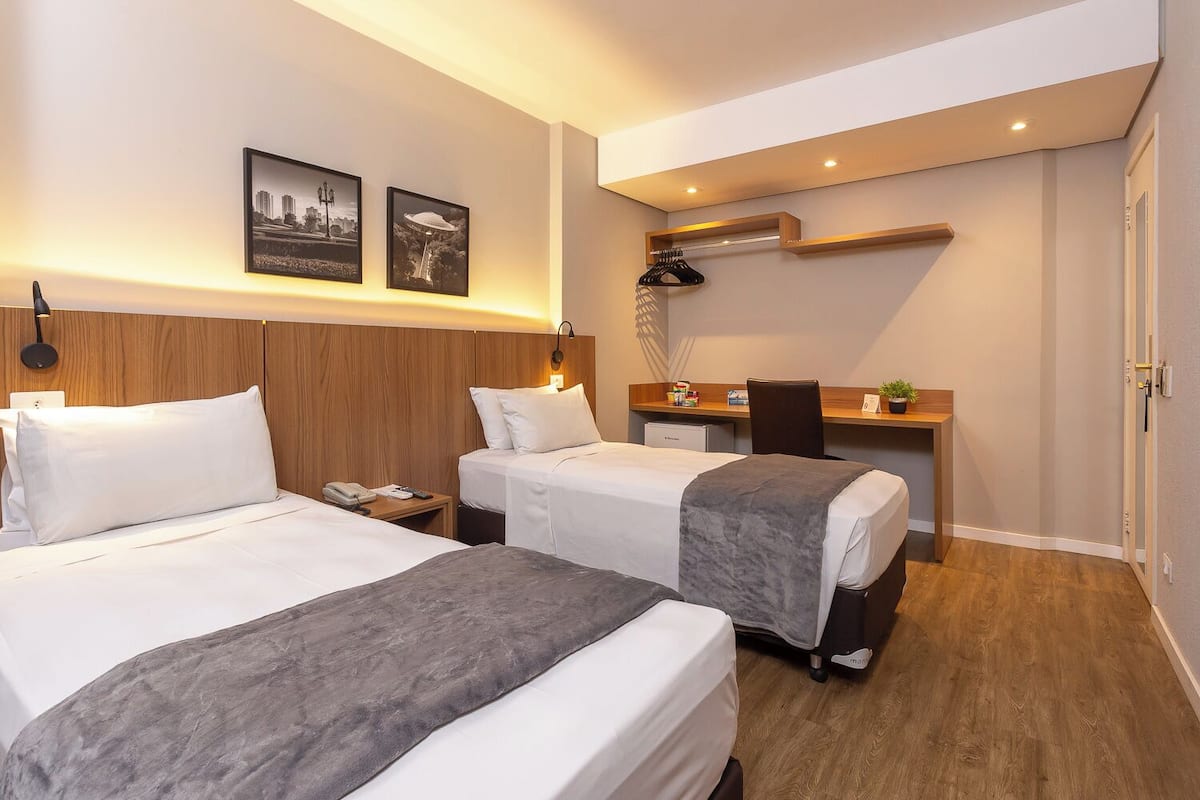 A hotel room with two beds, a desk, a chair, a small table, a potted plant, and a wall-mounted shelf.