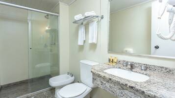 Un baño con ducha de vidrio, dos inodoros, un lavabo con encimera de granito y un secador de pelo montado en la pared.